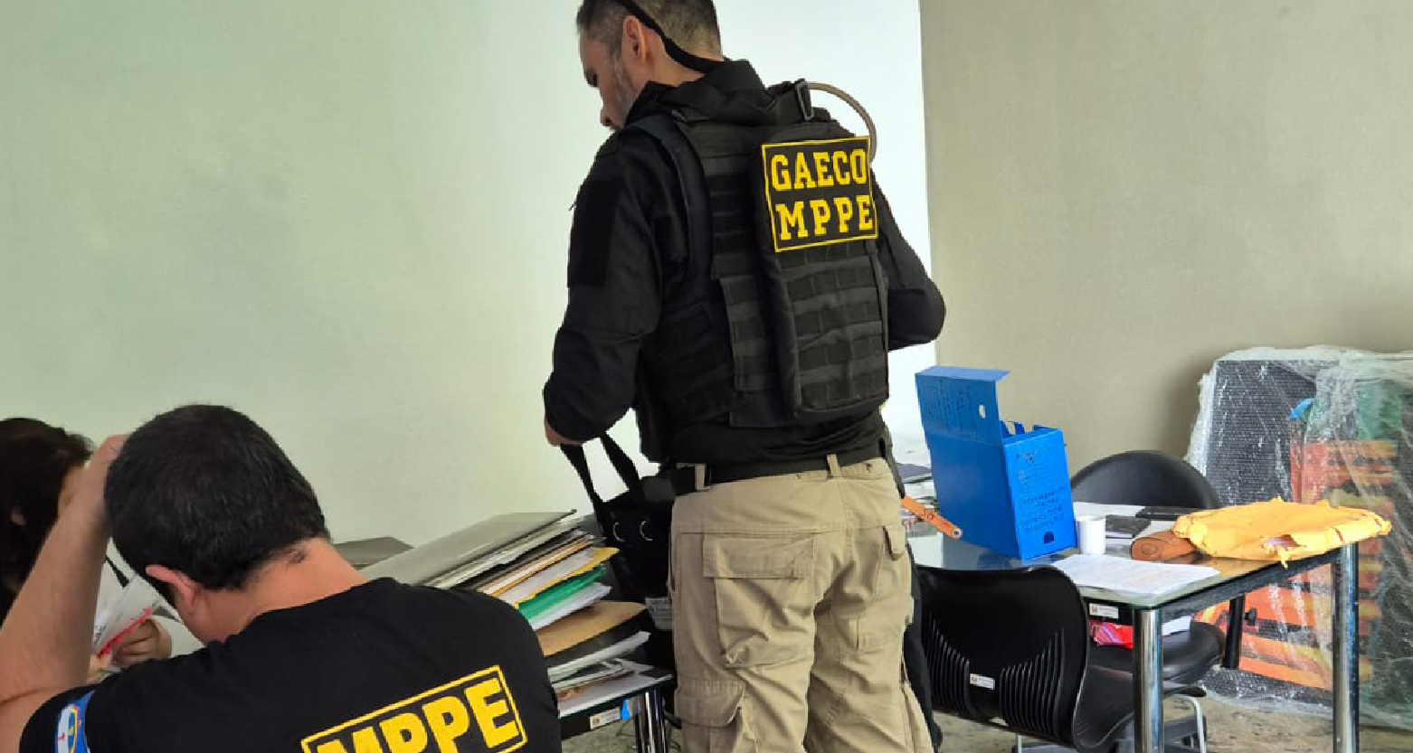 Um agente de colete com a inscrição "GAECO MPPE" vasculha documentos sobre uma mesa. Outro agente está sentado, olhando papéis.