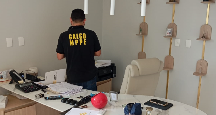 Pessoa com camisa preta com inscrição GAECO/MPPE busca documentos em um escritório