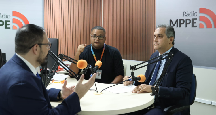 Três homens estão sentados em um estúdio de rádio. Um de óculos fala, gesticulando. Outro, à direita, escuta. No centro, um homem com camiseta preta e crachá observa.