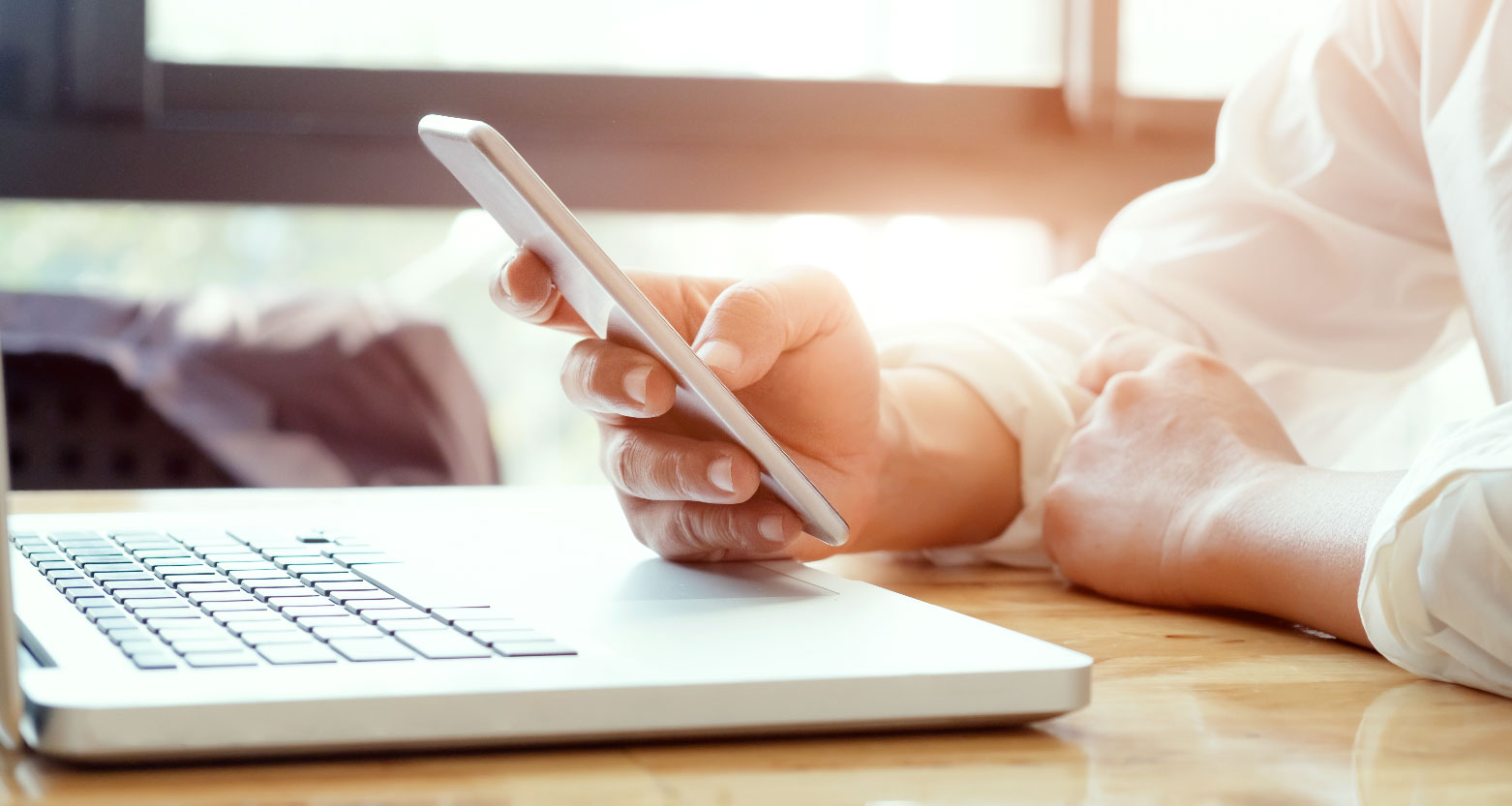 Fotografia de pessoa digitando em celular diante de notebook